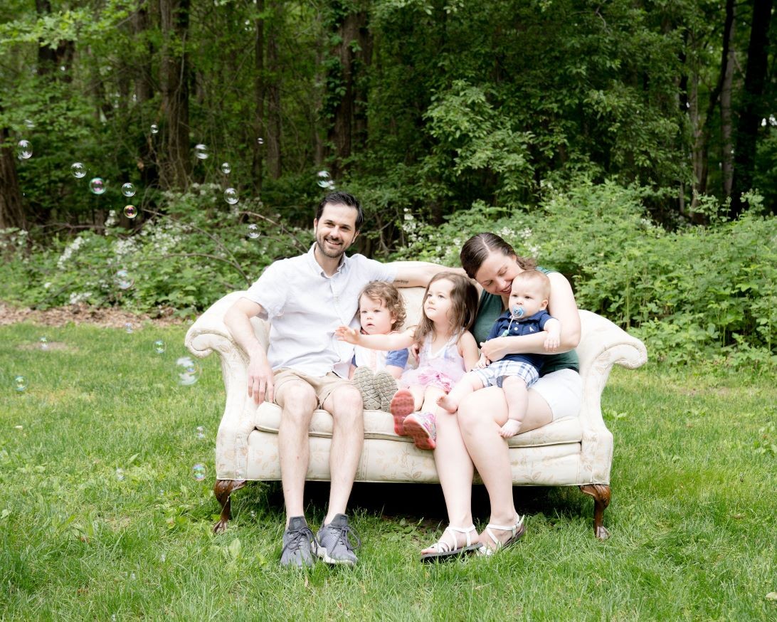 husband, wife and 3 young children sitting on a sofa together smiling and looking at bubbles in the air